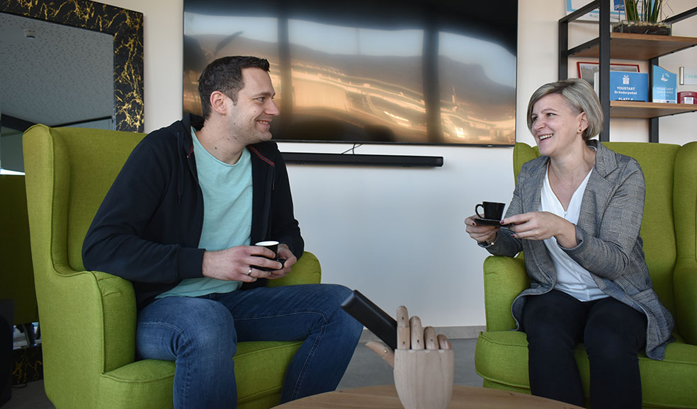 Zwei GTL Mitarbeiter sitzen im Foyer der GTL auf grüne Ohrensessel, trinken Kaffee und unterhalten sich